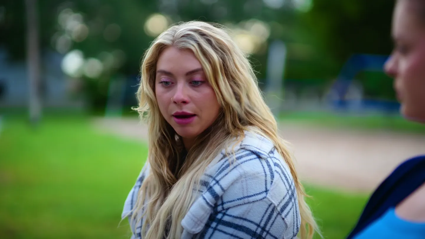 A young woman with long blonde hair, mid-sentence, the park soft behind her.