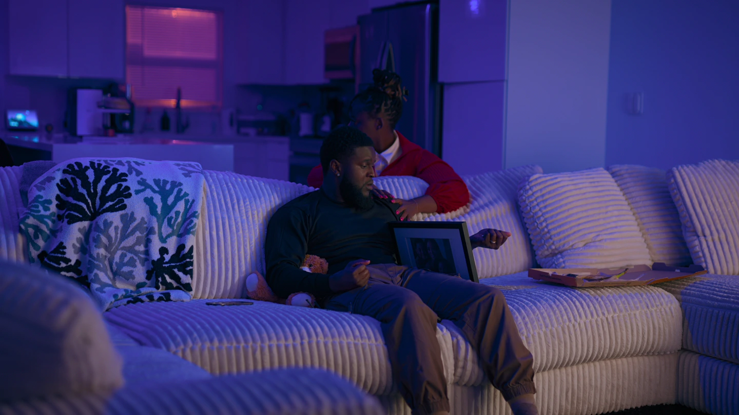 A man alone on a pale sectional sofa at night, a framed photograph propped beside him, the room washed in purple.