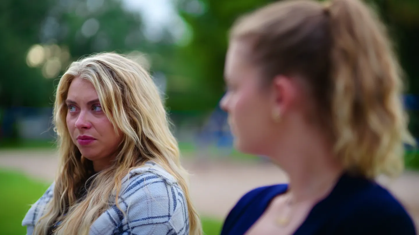 Over her sister's shoulder: the blonde woman meets her eye.