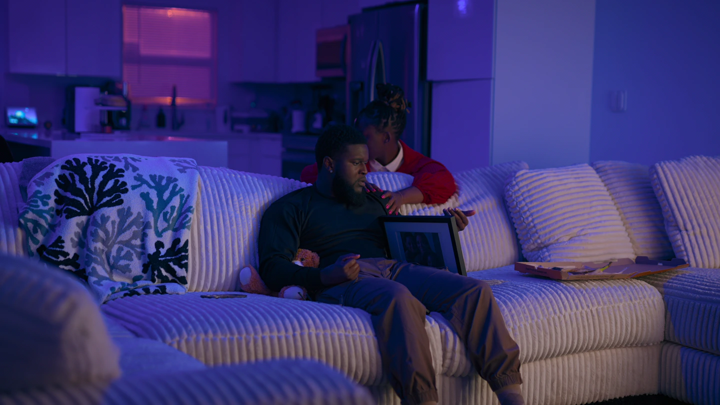 The same room from across the sofa, a woman in red standing behind him in the kitchen light.