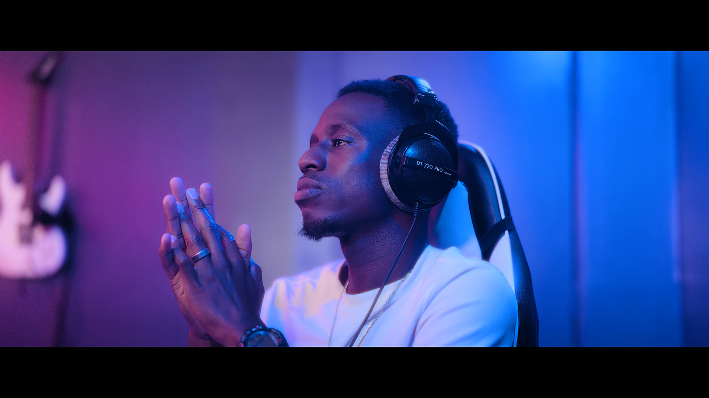 A man in studio headphones, hands pressed together, under magenta and blue light.