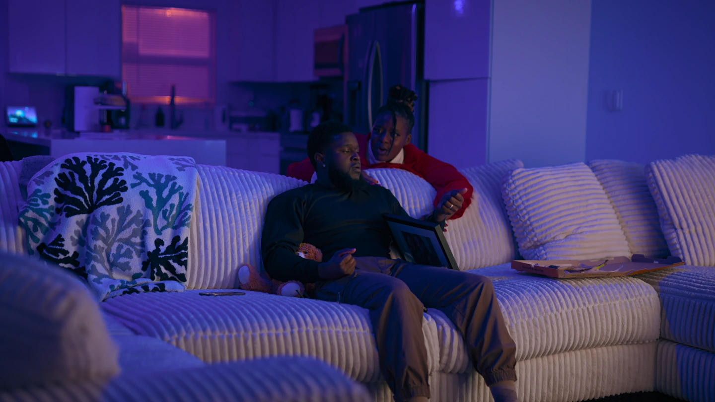 Wide of the living room, the woman close behind him as he sits forward.