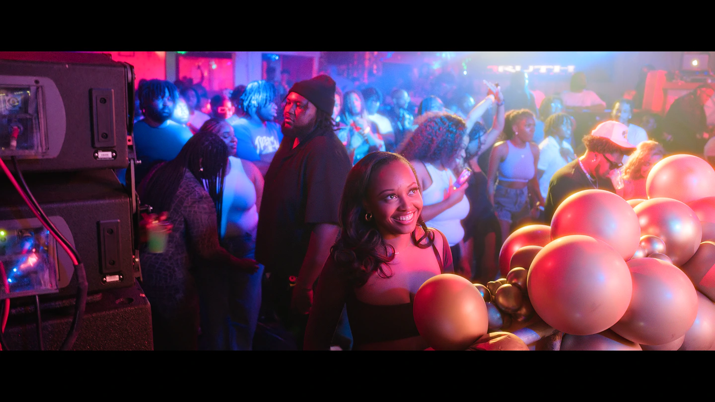 A packed party under red and blue light, balloons crowding the edge of the frame.